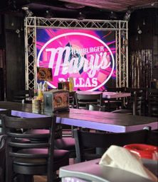 Vibrant Dallas burger bar interior with dark wooden tables and chairs, condiment caddies and napkins on communal tables, and a glowing pink circular stage sign framed by metal truss.