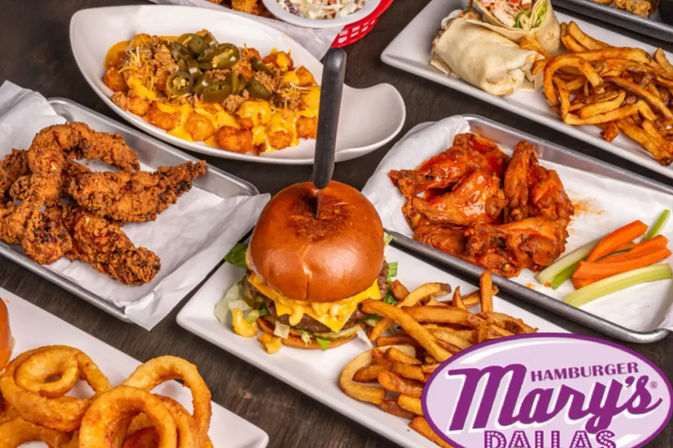 Close-up restaurant tabletop of American comfort food: cheeseburger with fries, buffalo wings with celery and carrots, crispy chicken tenders, loaded jalapeño tater tots, onion rings and a wrap on metal trays.