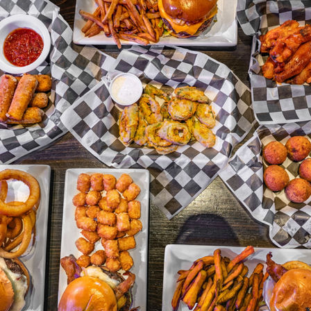 Top-down view of American pub food on a dark wooden table — cheeseburgers, sweet potato fries, onion rings, tater tots, mozzarella sticks, fried pickles, hushpuppies and buffalo wings on black-and-white checkered paper with dipping sauces.