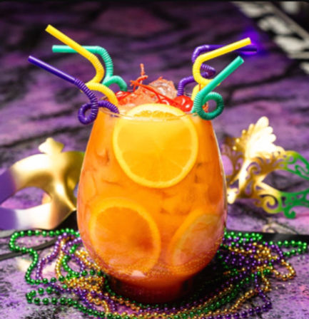 Festive Mardi Gras cocktail in a glass with orange slices and cherries over crushed ice, topped with colorful curly straws and surrounded by purple, green and gold beads and decorative masks on a purple backdrop.