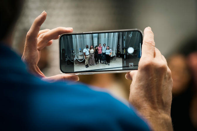 Someone snapping a smartphone group photo of six people posing in front of shiny stainless-steel fermentation tanks inside a craft brewery.