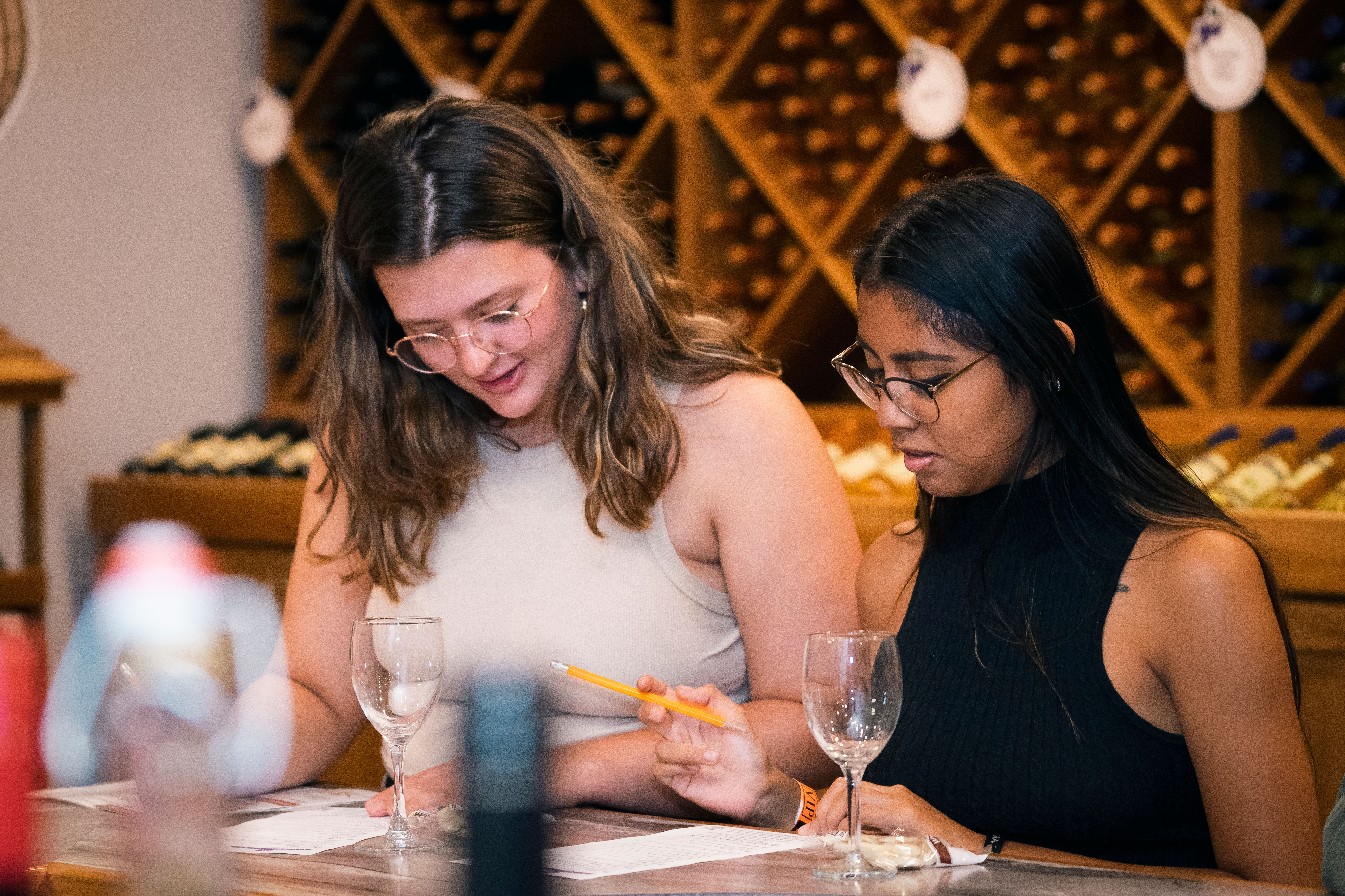 Two women at a cozy wine tasting room comparing tasting notes with a pencil and wine glasses, wine bottle racks lining the background.