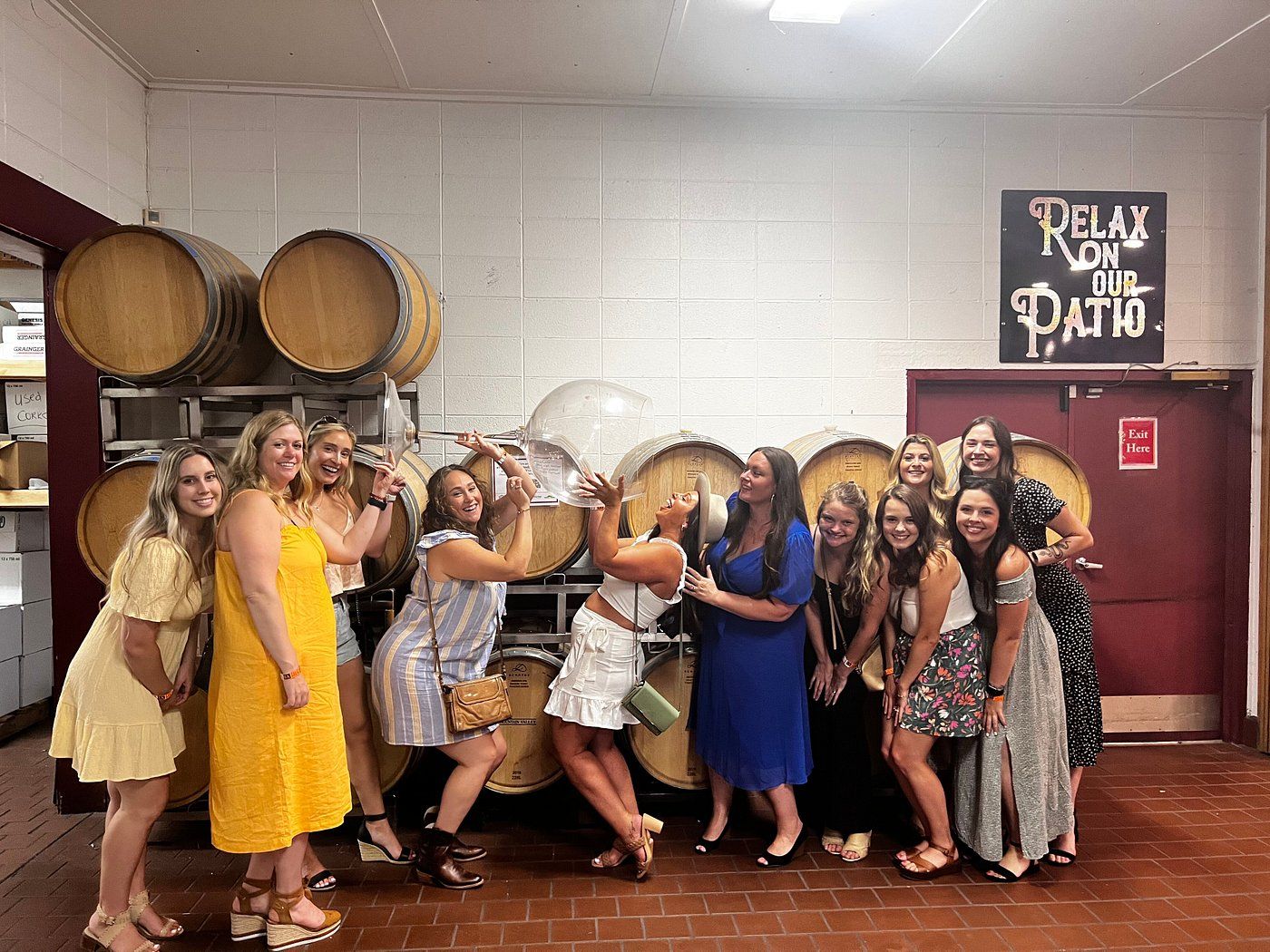Group of women in summer dresses laughing and posing inside a winery tasting room in front of stacked oak wine barrels, playfully using oversized clear wine-glass props beneath a 'Relax on our patio' sign.