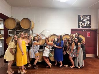 Group of women in summer dresses laughing and posing inside a winery tasting room in front of stacked oak wine barrels, playfully using oversized clear wine-glass props beneath a 'Relax on our patio' sign.