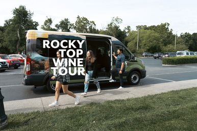 Three people stepping out of a green wine-tour van with bold white lettering, parked in a tree-lined parking lot on a sunny day, ready to explore local wineries.
