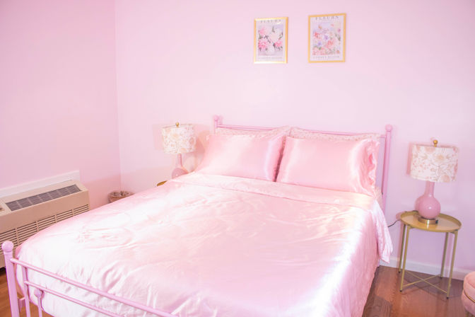 Bright pastel pink bedroom with a pink metal bed dressed in shiny satin comforter and matching pillows, pink walls, two floral bedside lamps on gold tables, framed floral prints above the bed and wood flooring.
