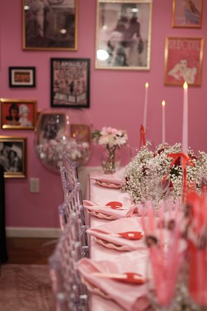 Pink-themed party table with blush napkins and red heart place tags, clear acrylic chairs, glassware, baby's breath floral centerpiece and lit pink taper candles against a gallery wall of framed photos.