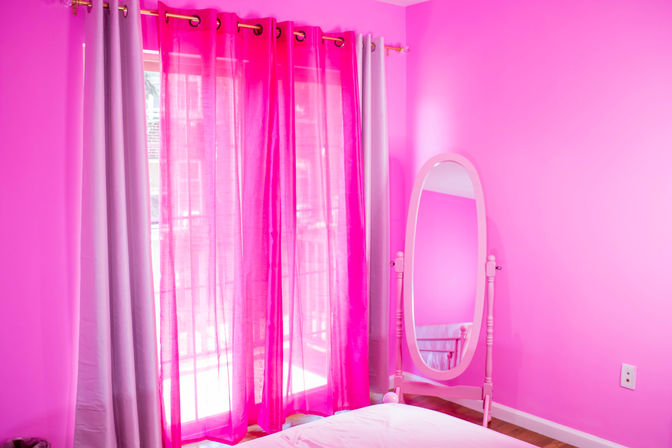 Vibrant pink bedroom interior with sheer fuchsia curtains over a sunlit window, lavender drapes, an oval standing mirror and bed corner on hardwood floor