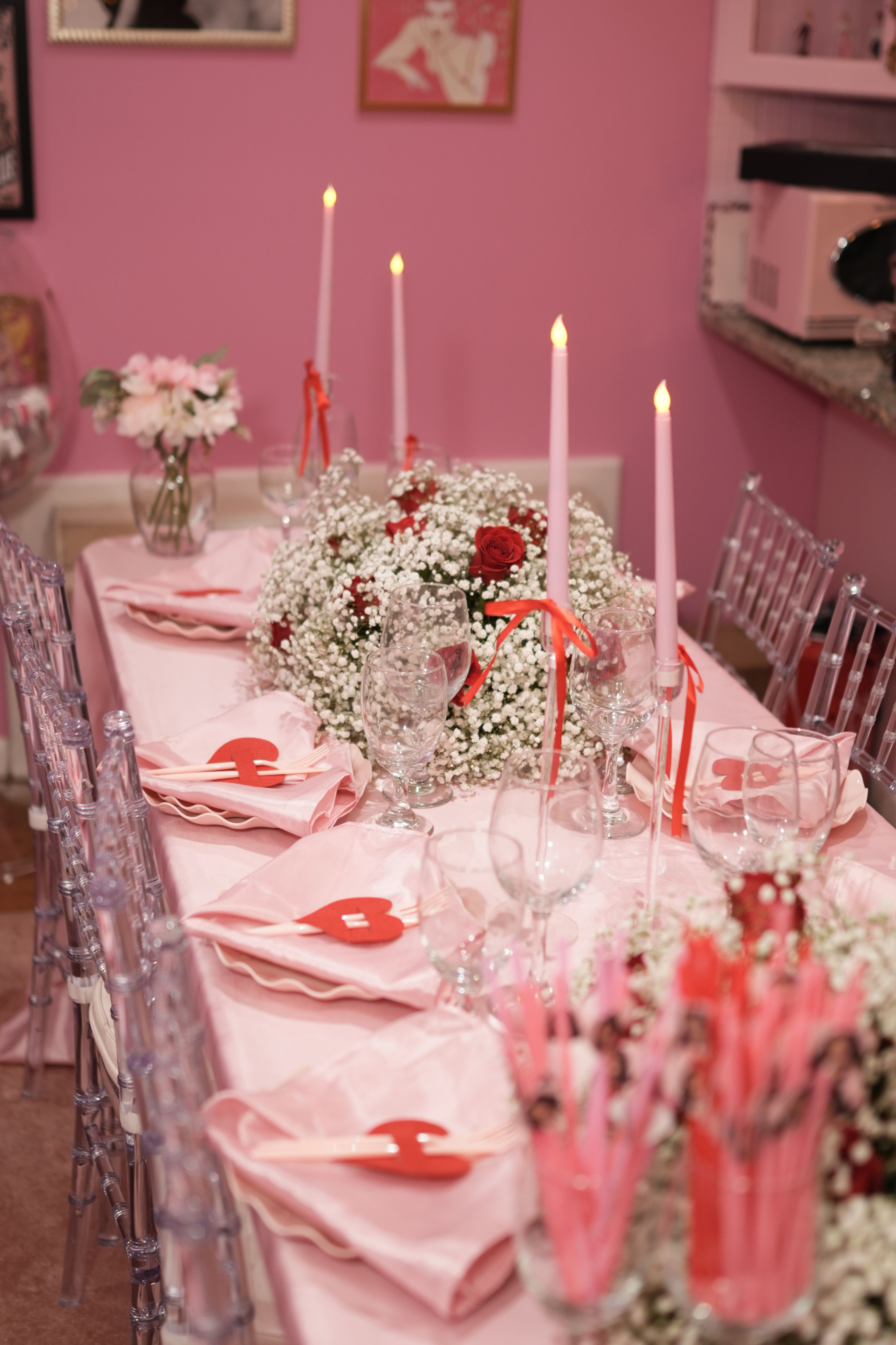 Pink-themed indoor romantic dining table setup with baby's-breath and red rose centerpiece, tall pink taper candles, clear glassware and folded pink napkins topped with red heart tags — Valentine-style party table decor.