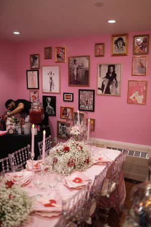 Pink party room with a gallery wall of vintage pin-up prints, long table dressed in pink linens, baby's-breath and red-rose centerpieces, tall pink taper candles, clear ghost chairs, and a server preparing drinks at a beverage station.