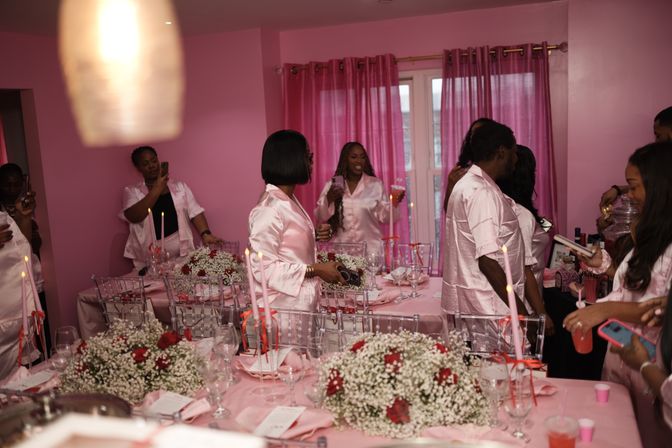Pink-themed indoor party with guests in matching pink satin robes mingling around tables set with pink linens, baby's-breath and red rose centerpieces, tall pink candles, glassware and drinks.