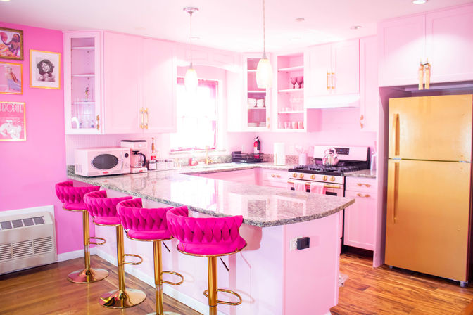 Bubblegum-pink kitchen with a large granite-look island, four hot-pink tufted bar stools on gold bases, pink cabinets and open shelving, gold refrigerator, range and pendant lights over hardwood floors.