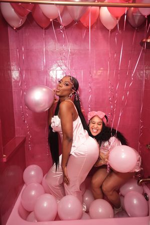 Two friends in pink satin pajamas laughing and posing in a pink-tiled bathtub filled with pink balloons and hanging ribbon streamers for a playful party vibe.