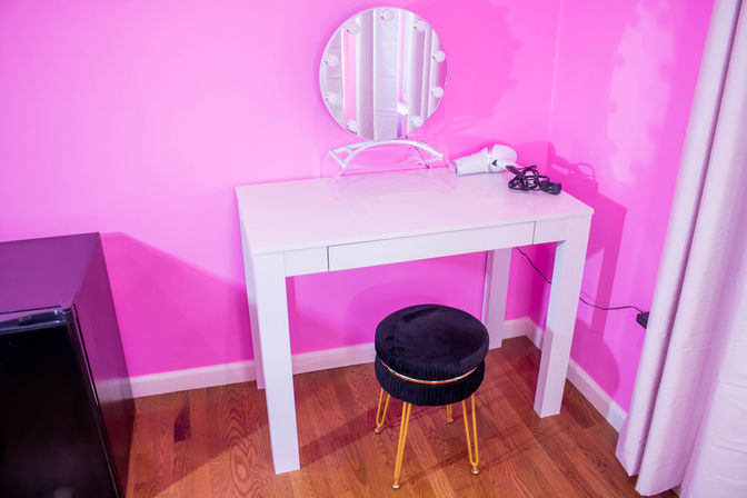 Cheerful pink dressing area with a white makeup vanity, round lighted mirror, clear acrylic stand, hair dryer and cord on top, black velvet stool with gold legs on hardwood floor.