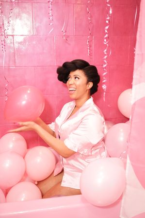 Smiling woman in a pink satin robe sitting in a bathtub filled with pastel pink balloons, holding a pink balloon and looking up against a glossy pink tiled backdrop with curling ribbon streamers.