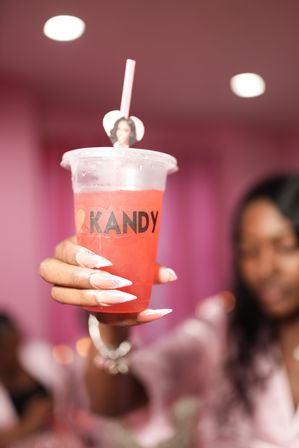 Close-up of a hand with long pink ombré nails holding a chilled pink beverage in a clear plastic cup with a heart stirrer, set against a blurred pink-themed indoor party background.