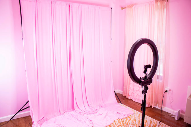 Pink home photography studio with floor-to-ceiling pink fabric backdrop on a stand, ring light on a tripod in front, matching pink walls and curtains, hardwood floor and patterned rug — ready for content creation.
