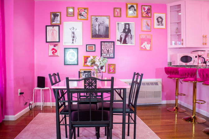Vibrant pink apartment dining nook with a black metal table and chairs, gallery wall of framed vintage photos, a vase of flowers on the table, and a kitchenette with plush pink bar stools and gold accents.