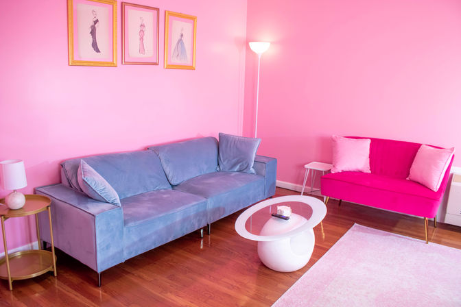 Bright pink living room with a blue velvet three-seat sofa, hot-pink loveseat with pale throw pillows, round white sculptural coffee table, gold side table, floor lamp, framed fashion sketches and hardwood floors.