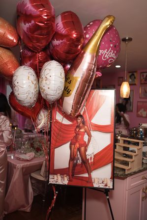 Indoor pink-themed birthday party display with red, white and gold balloons, a champagne-shaped balloon and a framed portrait of a woman in a red corset against draped backdrop, with table settings and pendant lights in the background.
