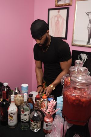 Person in a black shirt preparing drinks at an indoor pink-walled party drink station with assorted liquor bottles, glass dispensers of red punch and striped paper straws.