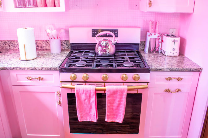 Bright pink home kitchen featuring a gas range oven with a patterned pink kettle on the stovetop, matching pink toaster, granite countertops, gold cabinet hardware, and two pink towels hanging on the oven handle.