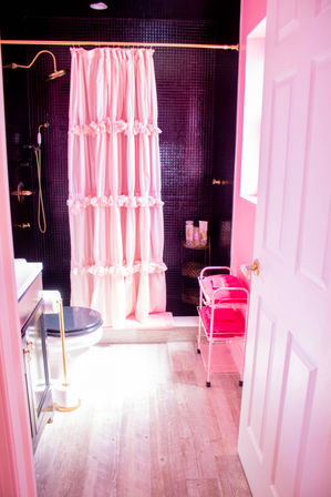 Bright pink bathroom with ruffled pink shower curtain over glossy black tile shower, brass fixtures, black toilet seat, light wood plank floor and pink towel rack with folded towels.