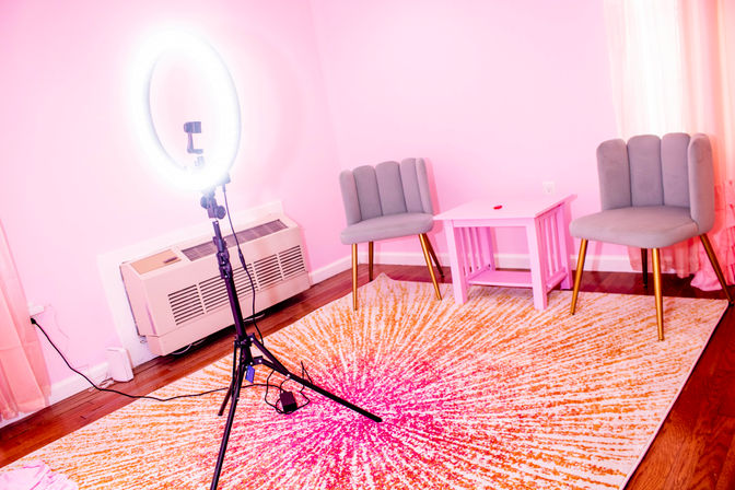 Bright pink content studio with an illuminated ring light on a tripod, two gray scalloped chairs with gold legs, a small pink side table, and a vibrant pink-and-orange sunburst rug on hardwood floor.