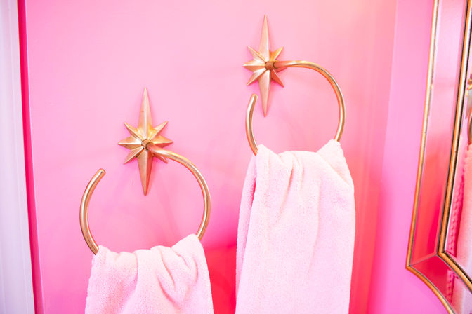 Cheerful hot-pink bathroom wall with two gold star-shaped towel rings holding plush pale-pink towels, mirrored trim visible at the edge