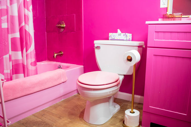 Vibrant pink residential bathroom featuring a white toilet with a pink seat, pink bathtub with patterned pink shower curtain, matching pink vanity, gold-tone fixtures and wood-look flooring.