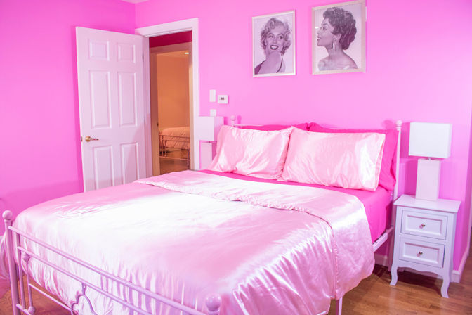 Vibrant pink bedroom with shiny satin pink bedding on a white metal bed frame, white nightstand and lamp, hardwood floor, open door and retro black-and-white glamour portraits on the wall.