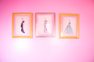 Three gold-framed vintage fashion illustrations of elegant gowns displayed on a bright pink wall — playful retro wall art.