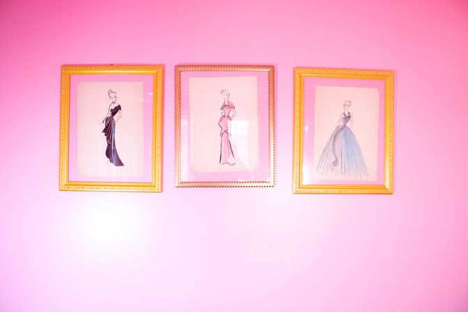 Three gold-framed vintage fashion illustrations of elegant gowns displayed on a bright pink wall — playful retro wall art.