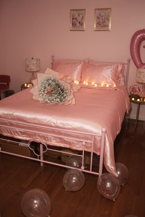 Cozy pink bedroom with satin blush bedding on a metal bed frame, a bouquet made from folded banknotes on the bed, lit tea candles along the headboard and clear confetti balloons on the hardwood floor.