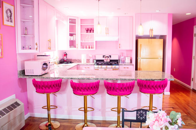 Bright pink kitchen with a granite island and four tufted fuchsia bar stools with gold bases, white cabinets with open shelving, gas range, yellow refrigerator, pendant lights and hardwood floors.