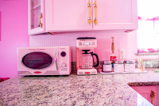 Retro pink kitchen counter with a pink microwave and coffee maker, labeled coffee, creamer and sugar canisters, gold cabinet hardware and granite countertop