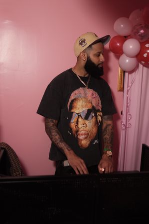 Bearded man in a tan cap and graphic T-shirt stands against a pink wall with heart balloons, looking sideways while working at a DJ booth during an indoor party.