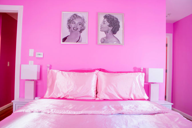 Playful bright pink bedroom interior with satin pink bedding, two white bedside lamps, and framed vintage celebrity portraits above the headboard, stylish home decor scene.