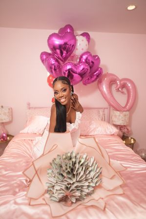 Smiling woman lounging on pink satin bedding in a playful pink bedroom photoshoot, surrounded by heart-shaped balloons and a bouquet made of folded cash.
