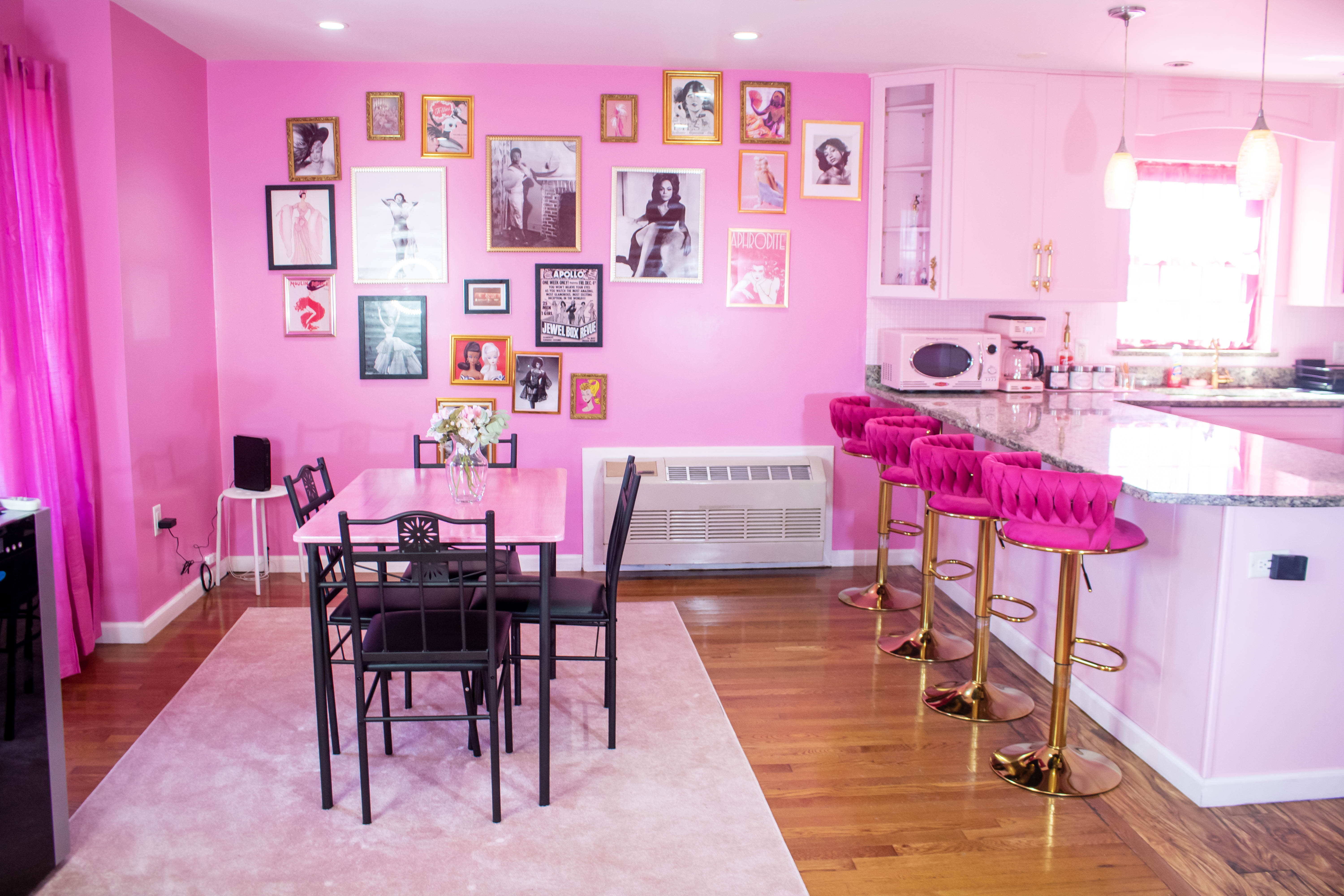 Open-plan bright pink dining and kitchen with a black metal dining table, gallery wall of framed vintage photos, marble breakfast bar with hot-pink tufted velvet bar stools on gold bases, hardwood floors and pink curtains.