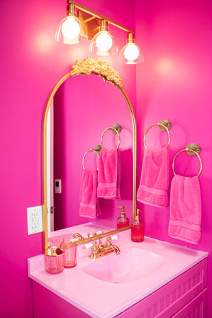 Vibrant hot-pink powder room vanity with gold arched mirror and fixtures, matching pink towels, soap dispenser and glass cups.