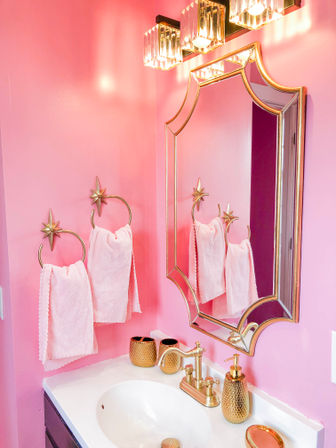 Vibrant pink bathroom vanity with white sink, gold faucet and ornate gold-framed mirror, star-shaped towel rings holding soft pink towels, crystal light fixture and matching gold soap dispenser and accessories.