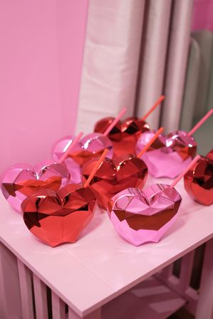 Playful shiny faceted heart-shaped metallic cups in red and pink with matching straws clustered on a pink table against a pink backdrop