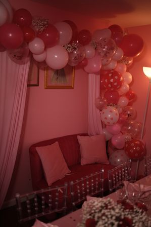 Indoor Valentine's-themed party corner with pink and red balloon garland and confetti balloons framing a cozy pink loveseat with pillows, sheer curtains and soft lamp lighting — romantic event decor.