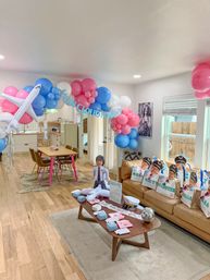 Airplane-themed baby shower in a bright home living room with pink, blue and white balloon arch and inflatable plane, 'On Cloud Nine' banner, pilot cutout, pilot-hat goodie bags on a tan sofa, and a coffee table of themed favors.