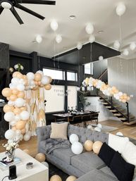 Open-concept modern living room and dining area decorated for a bridal shower with white and beige balloon garlands, helium balloons on the ceiling, a balloon arch by the windows, set dining table, gray sectional sofa and floating staircase.