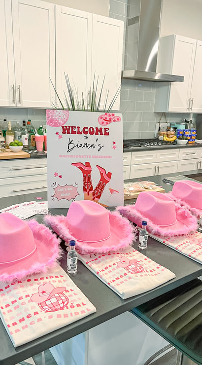 Modern white kitchen island styled for a bachelorette weekend with pink feather-trim cowboy hats, folded printed tees, tiny liquor bottles, and a festive welcome sign.