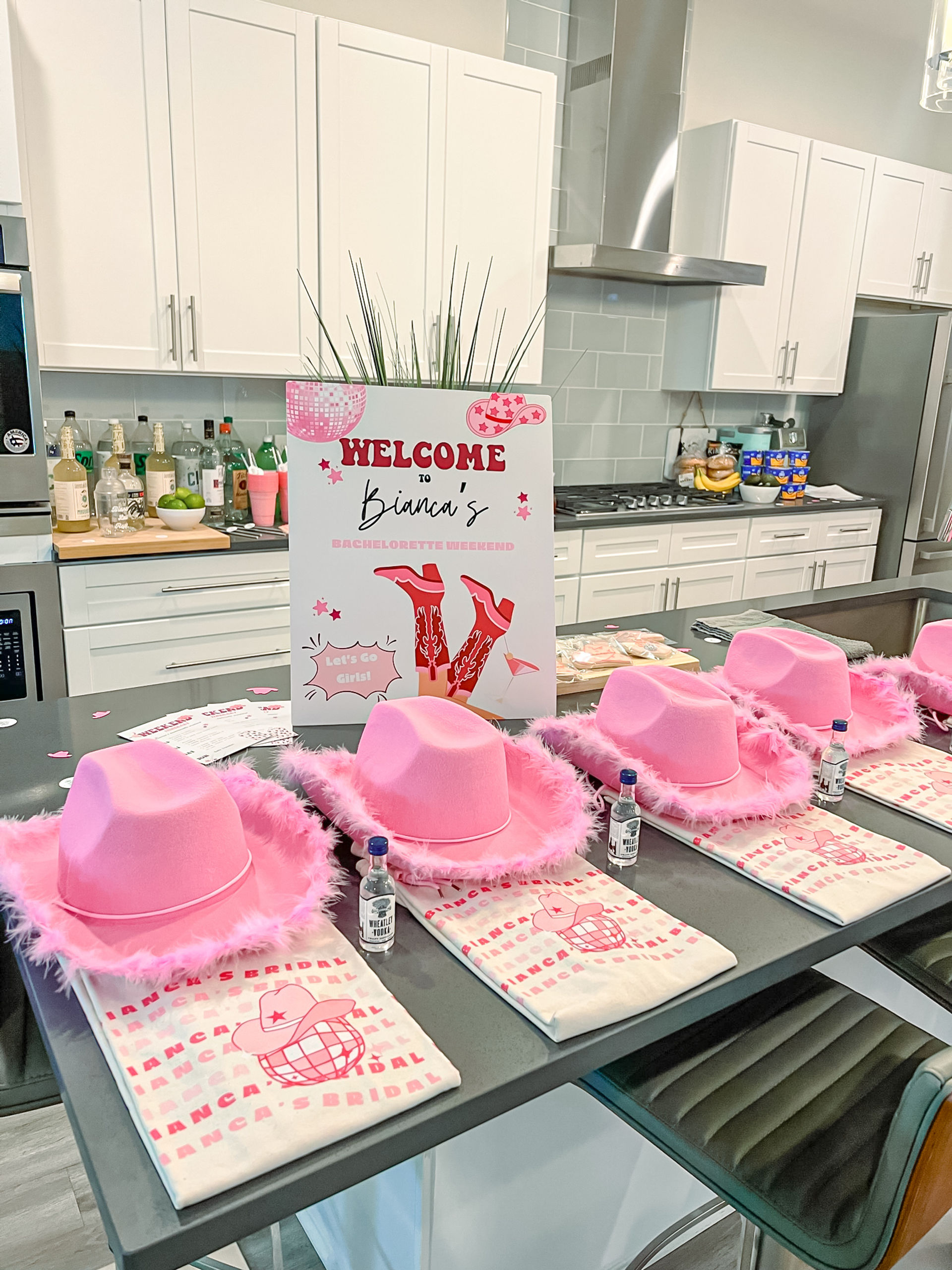 Modern white kitchen island styled for a bachelorette weekend with pink feather-trim cowboy hats, folded printed tees, tiny liquor bottles, and a festive welcome sign.