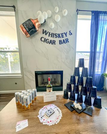 Living-room whiskey and cigar bar party setup: whiskey-bottle balloon and white balloons over a marble fireplace with a 'Whiskey & Cigar Bar' banner; table with a whiskey bottle, cans of beer, fan of playing cards, stacked black plastic cups and ping-pong balls.