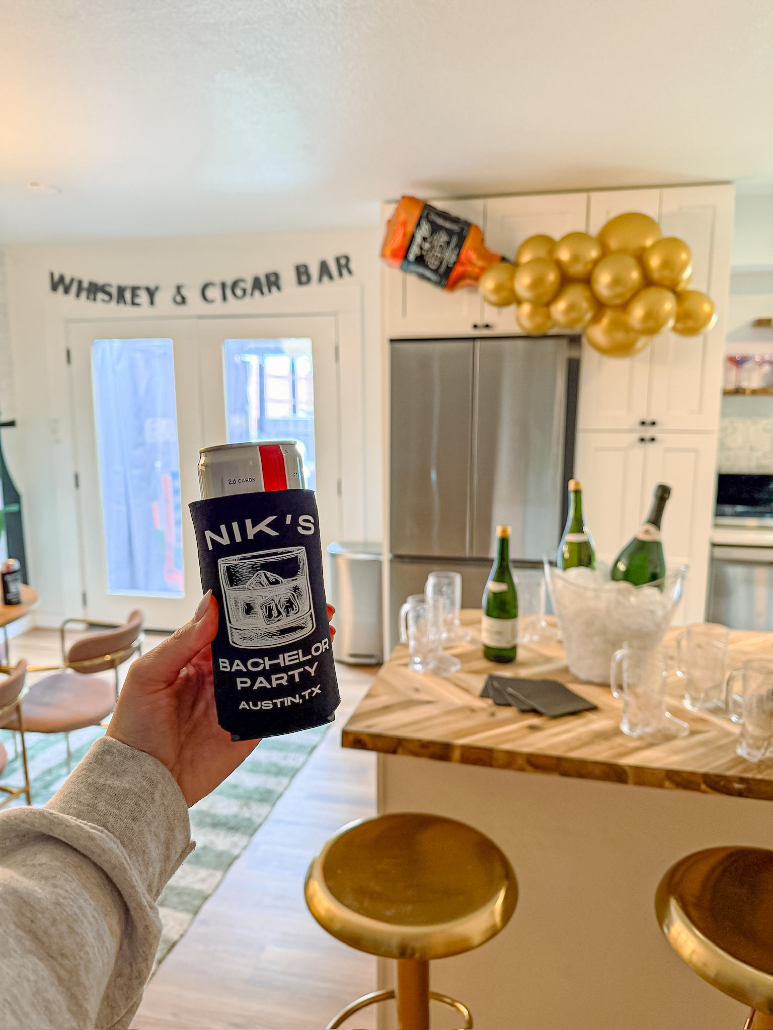 Hand holding a canned drink in a bachelor-party koozie (Austin, TX) in a kitchen with champagne bottles, a "Whiskey & Cigar Bar" banner and a cluster of gold balloons.